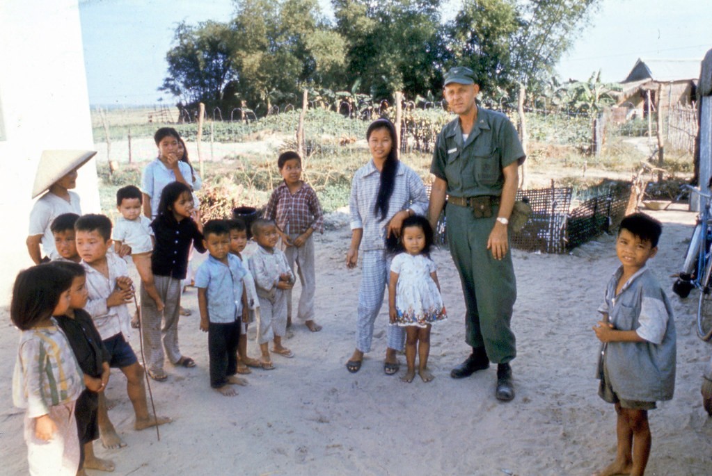 The young lady with the long black hair, is a senior in high school at Danang. Her brother in the checkered shirt on her right accompanies her as she acts as an interpreter between the dental officer and the Vietnamese people as he makes his rounds of the camps and villages.