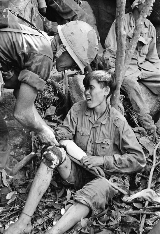 Medical Spec. 4 Roy L. Johnson of Houston, Texas, treats the bloodied knee and arm of one of a group of captured North Vietnamese soldiers on a rocky cliff near Dong Tre, 225 miles northeast of Saigon, in June 1966