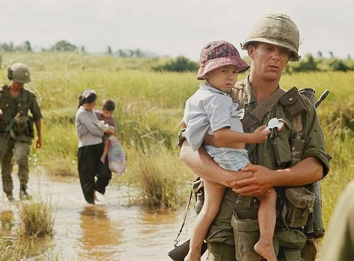 Một người lính Mỹ dù đang hành quân rất vất vả nhưng vẫn sẵn lòng bế theo một em bé Nam Việt Nam.